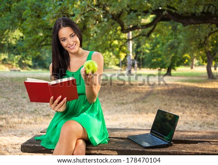 Beautiful young woman with toothy smile holding book and apple in the park and sitting on a wooden table beside laptop.