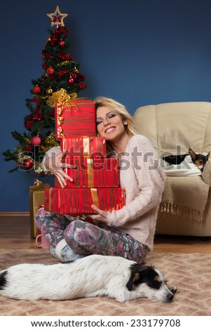 Beautiful young woman sitting near Christmas tree and holding a gifts in arms with the dog in front of her who sleeps on the floor.