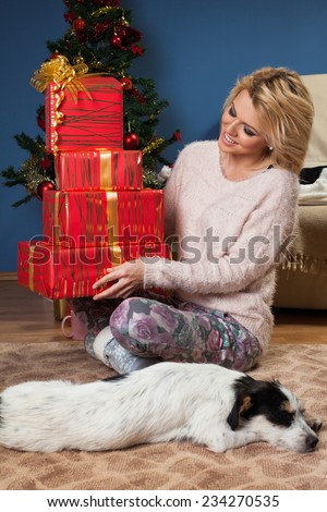 Beautiful young woman sitting near Christmas tree and holding a gifts in arms with the dog in front of her who sleeps on the floor.