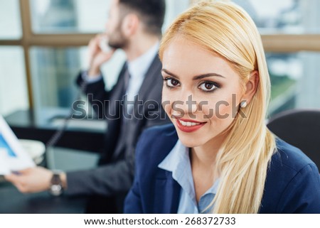 Beautiful young businesswoman posing with a smile in office with a man out of focus in the background