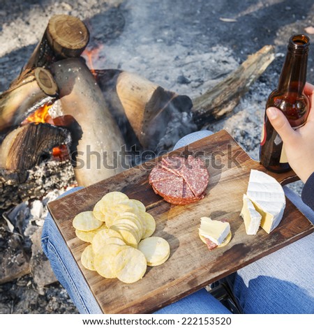 Gourmet camping food on a wooden board, with cheese, salami and crackers with a beer in front of the fire