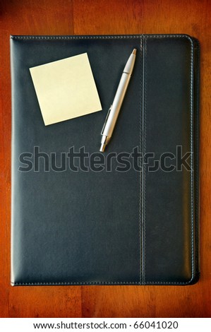 A black leather executive folder on timber desk with sticky note and silver pen