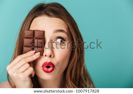 Portrait of a cheery brown haired woman with bright makeup holding chocolate bar at her face isolated over blue background