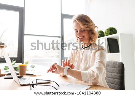 Image of cheerful young business woman sitting in office using laptop computer looking at watch.