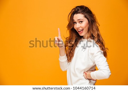 Happy brunette woman in sweater posing with arm on hip while having idea and looking at the camera over yellow background