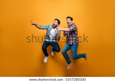 Full length portrait of a two excited young men pointing fingers away while jumping isolated over yellow background