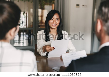 Business, career and recruitment concept - young asian woman smiling and holding resume while interviewing as candidate for job in big company