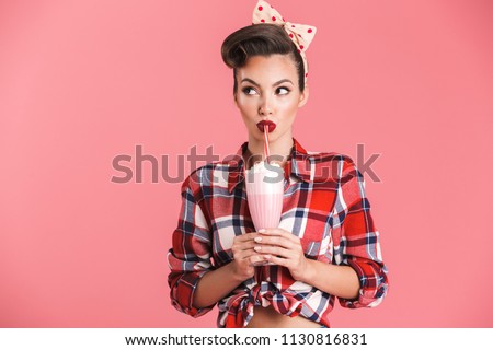 Portrait of a cheerful brunette pin-up girl in plaid shirt holding milk shake and looking away over pink background