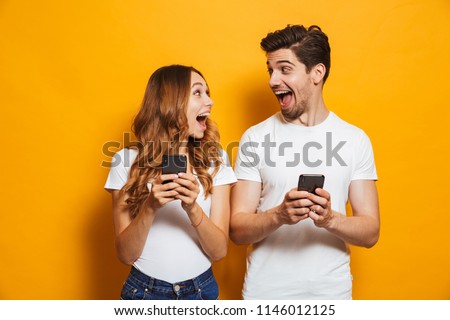 Photo of positive excited people man and woman screaming and looking at each other while both using mobile phones isolated over yellow background