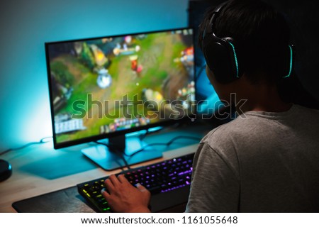 Image of immersed teenage gamer boy playing video games on computer in dark room wearing headphones and using backlit colorful keyboard