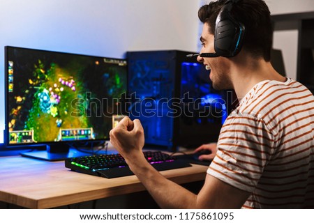 Portrait of delighted young guy playing video games on computer wearing headphones and using backlit colorful keyboard