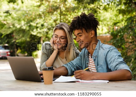 Happy young multiethnic couple spending time together at the park, studying while sitting at the table with laptop