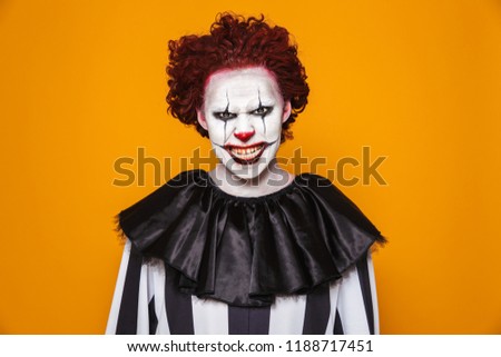 Angry clown man 20s wearing black costume and halloween makeup looking at camera isolated over yellow background