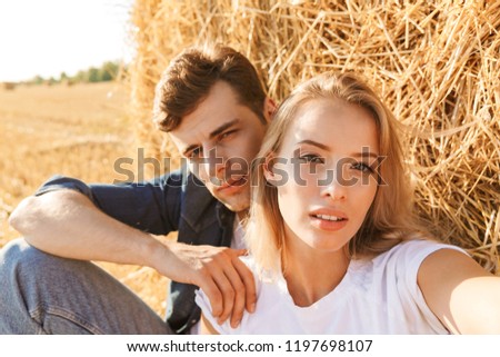 Photo of young couple man and woman taking selfie while sitting under big haystack in golden field during sunny day