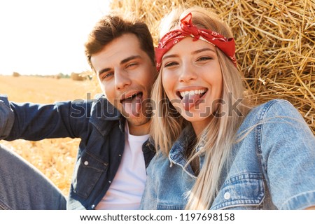Photo of smiling couple man and woman smiling while sitting under big haystack in golden field during sunny day