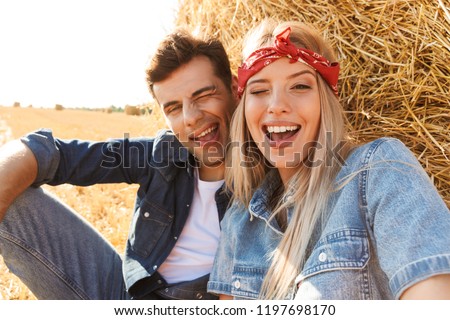 Photo of cute couple man and woman smiling while sitting under big haystack in golden field during sunny day