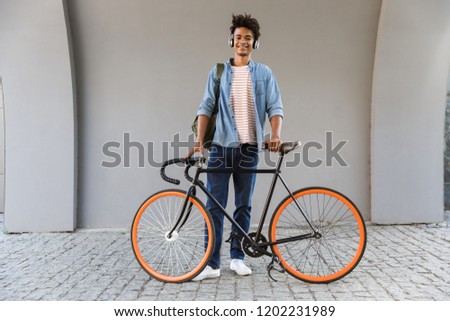 Smiling young african man outdoors, standing with bicycle, listening to music with headphones