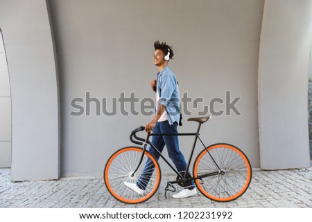Smiling young african man outdoors, walking with bicycle, listening to music with headphones