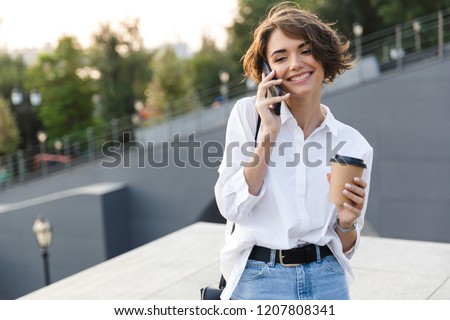 Attractive young woman standing at the street, holding cup of coffee, talking on mobile phone