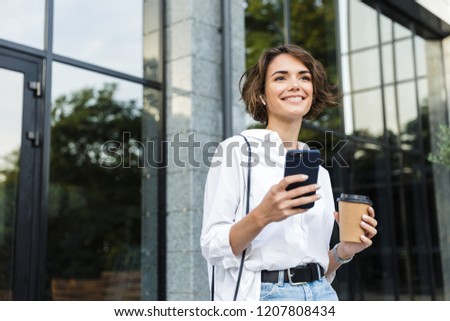 Beautiful young woman in earphones standing outdoors at the street, talking on mobile phone, drinking coffee