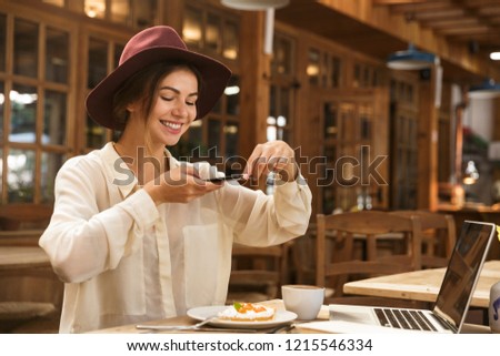 Cheerful young woman taking a picture of her lunch while sitting at the cafe table with laptop computer