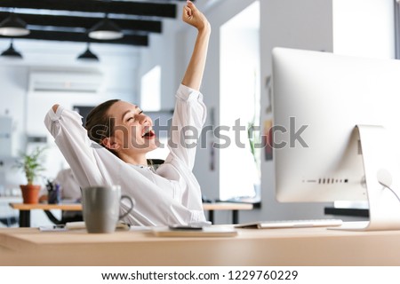Happy young woman dressed in shirt sitting at her workplace at the office, resting, stretching her hands