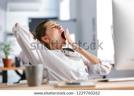 Sleepy young woman dressed in shirt sitting at her workplace at the office, yawning