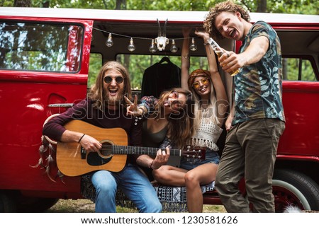 Group of joyous hippies happy men and women laughing and playing guitar near vintage minivan into the nature