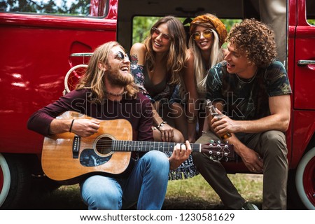 Group of young hippies men and women laughing and playing guitar near vintage minivan into the nature