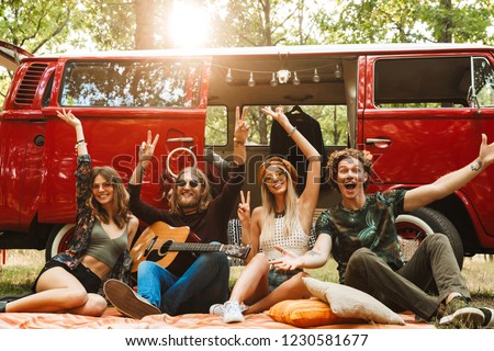 Group of young hippies men and women laughing and sitting near vintage minivan into the nature
