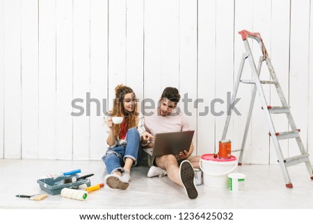 Photo of excited couple man and woman sitting on floor over white wall near ladder and using laptop while making renovation indoor