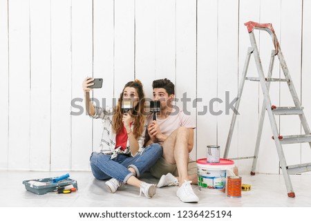 Photo of adorable couple man and woman sitting on floor over white wall near ladder and taking selfie on cell phone while making renovation indoor