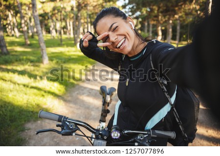 Attractive fit sportswoman riding on a bicycle at the park, listening to music with wireless earphones, taking a selfie