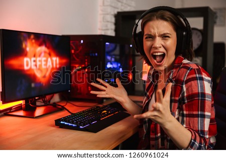 Angry girl gamer sitting at the table, playing online games on a computer indoors