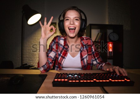 Excited girl gamer sitting at the table, playing online games on a computer indoors, celebrating success, showing ok