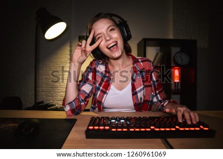 Excited girl gamer sitting at the table, playing online games on a computer indoors, celebrating success
