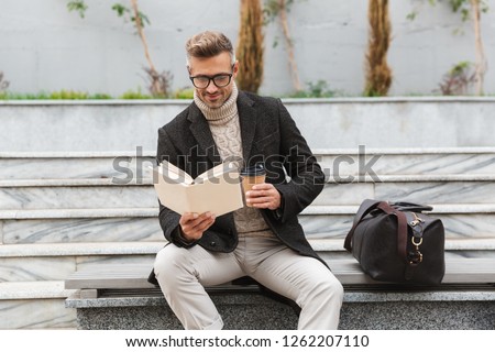 Handsome man wearing jacket reading a book while sitting outdoors