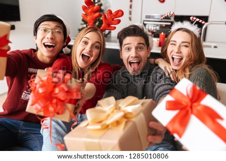 Group of happy young friends celebrating New Year together while sitting on a sofa at home, giving presents