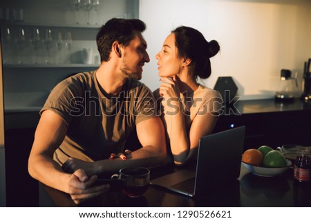 Beautiful young couple embracing while sitting at the kitchen table with laptop computer
