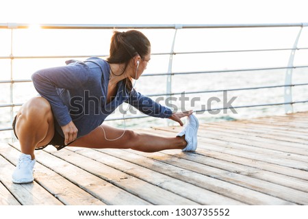 Image of a beautiful young sports fitness woman make stretching exercises at the beach outdoors listening music with earphones.