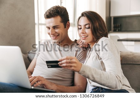 Smiling young couple relaxing on a couch at home, using laptop computer, shopping with credit card