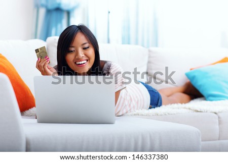 Happy asian woman shopping on-line while lying on the sofa in her living room