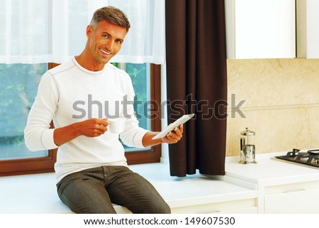Happy young man at home drinking coffee and reading news on his electronic tablet