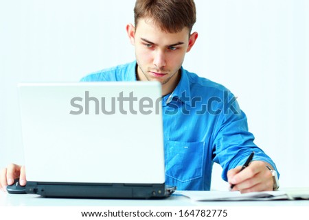 Young concentrated businessman writing notes down and looking at laptop in office