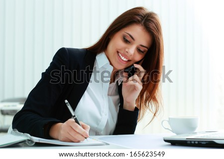 Young happy businesswoman talking on the phone and writing notes in office