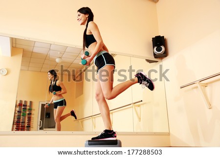 Young smiling fit woman doing exercises with dumbells on step board at gym
