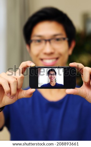 Smiling asian man taking self picture with smartphone camera. Focus on smartphone