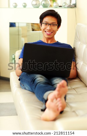 Young happy asian man lying on the sofa with laptop at home