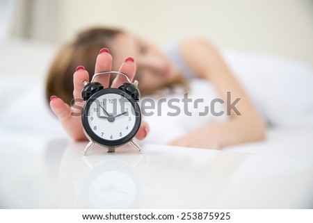 Sleepy woman in bed extending hand to alarm clock. Focus on clock