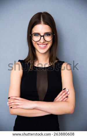 Portrait of a happy woman with arms folded and glasses standing over gray background. Wearing in fashion black dress. Looking at camera